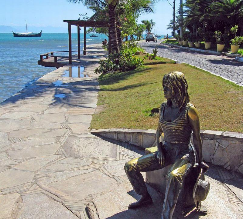 Estátua de bronze na orla Bardot em Búzios, com calçadão, mar e barcos ao fundo